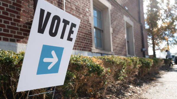 A white sign that says, "vote" in bold, black letters with a white arrow planted in a bush outside of a brick building.
