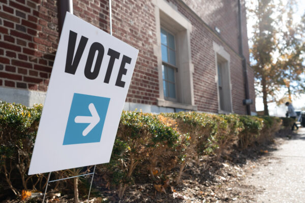 A white sign that says, "vote" in bold, black letters with a white arrow planted in a bush outside of a brick building.