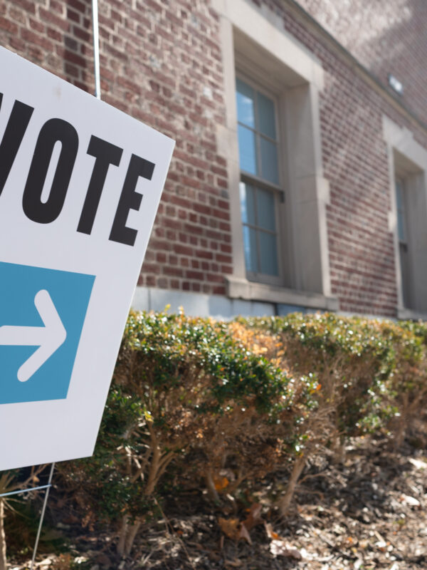 A white sign that says, "vote" in bold, black letters with a white arrow planted in a bush outside of a brick building.