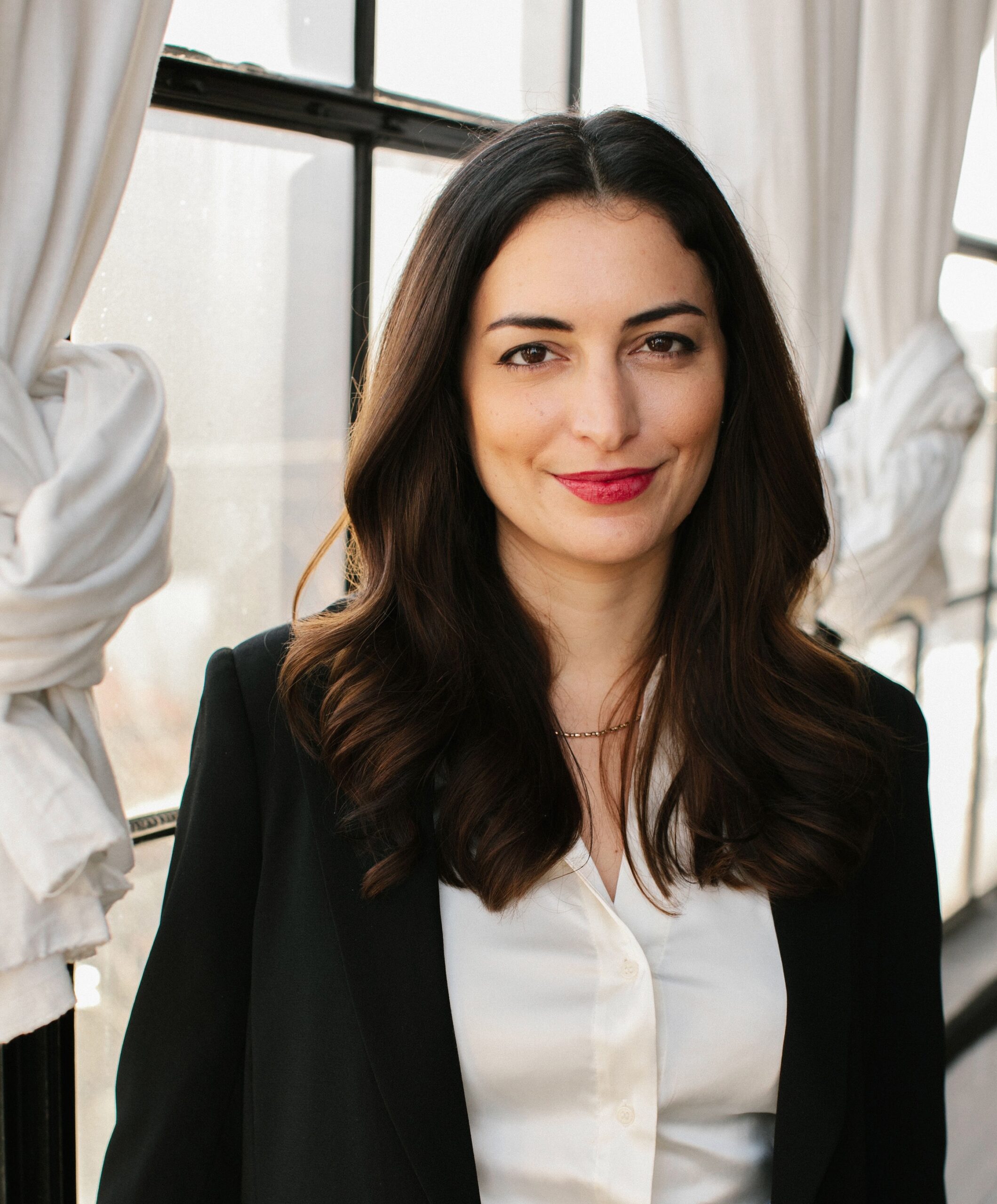 Kate smiling while wearing a white collared shirt and black blazer while in front of a window