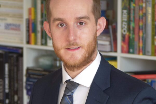 A picture of Keith Armstrong with a beard and navy blue suit with a white button down shirt with a blue plaid tie