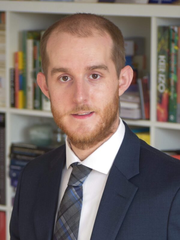 A picture of Keith Armstrong with a beard and navy blue suit with a white button down shirt with a blue plaid tie