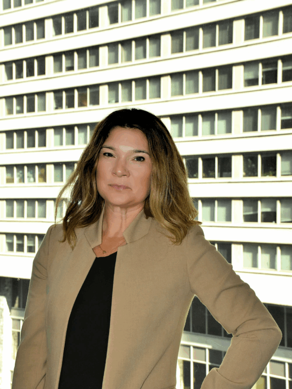 A picture of Rebecca in front of a window with a brown blazer, black shirt, and highlighted brunette and blonde hair.