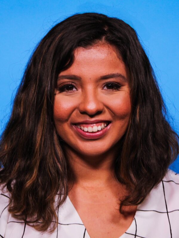 Jessica Riestra standing and smiling in front of a blue background in a white and black plaid blouse