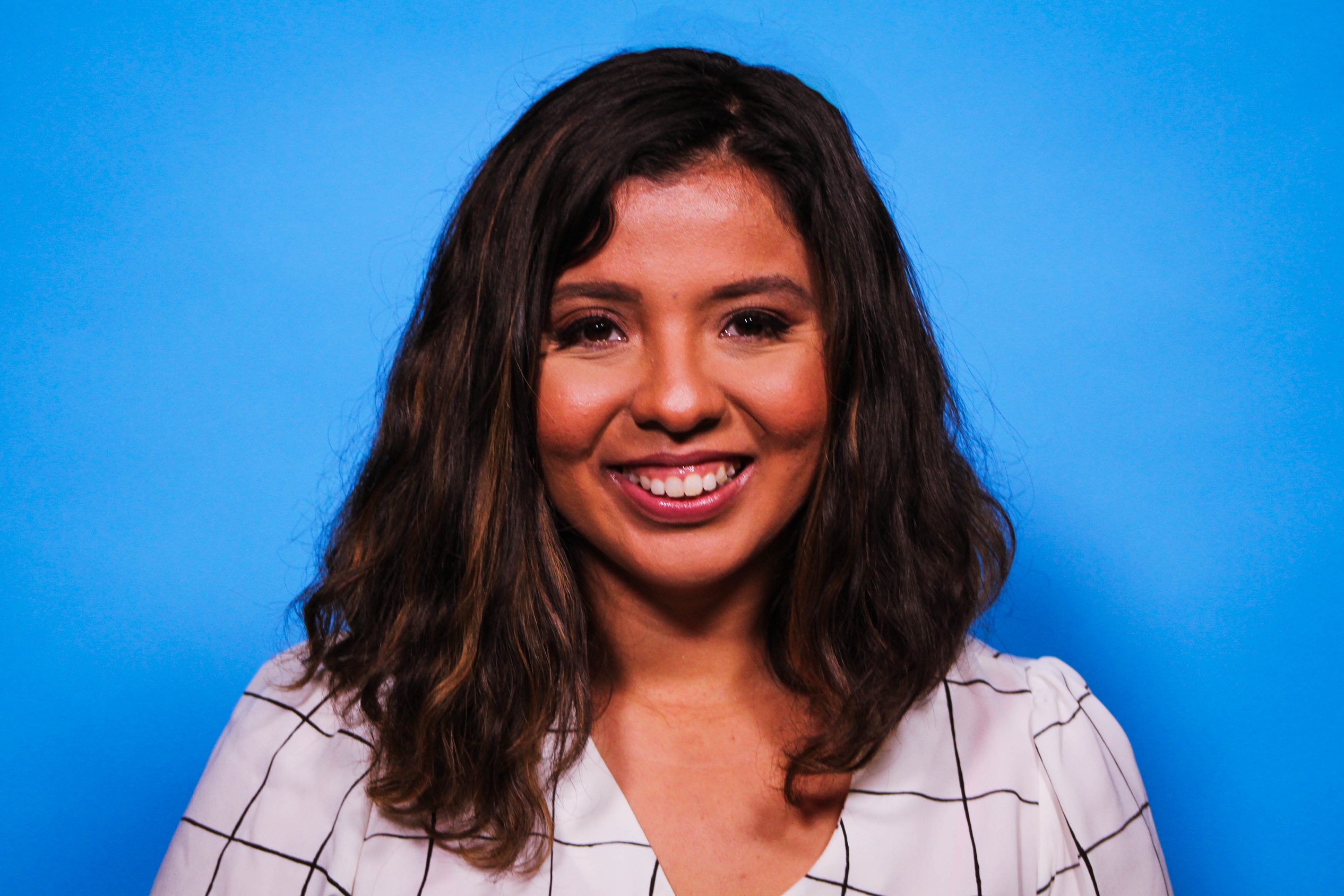 Jessica Riestra standing and smiling in front of a blue background in a white and black plaid blouse