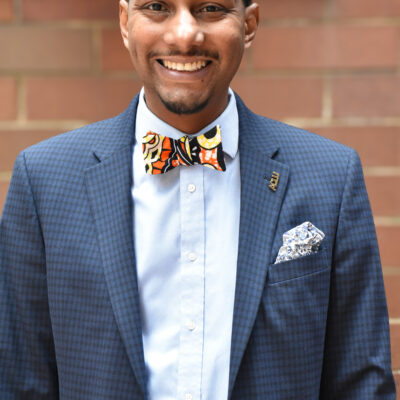 A photo of Mike Lee, smiling, with a light blue button down and dark blue suit blazer with a kente cloth bowtie.