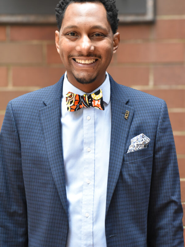 A photo of Mike Lee, smiling, with a light blue button down and dark blue suit blazer with a kente cloth bowtie.