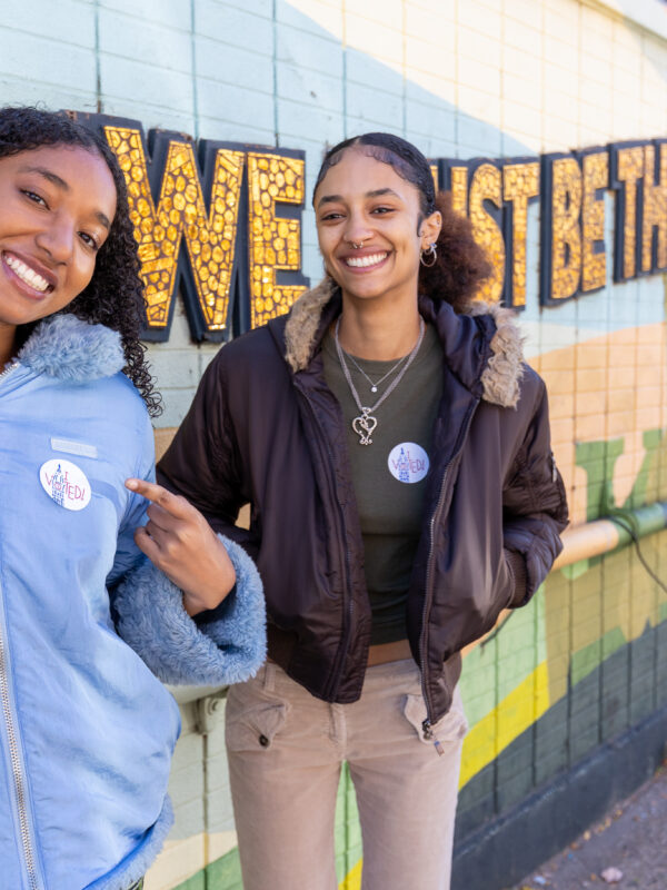 Two people, one in a blue puffer jacket and one in a brown coat pointing to their voting stickers on their lapels