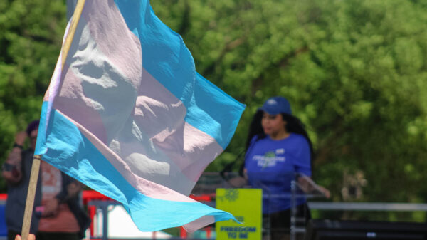 Trans pride flag with a woman speaking on stage in the background.