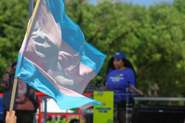 Trans pride flag with a woman speaking on stage in the background.