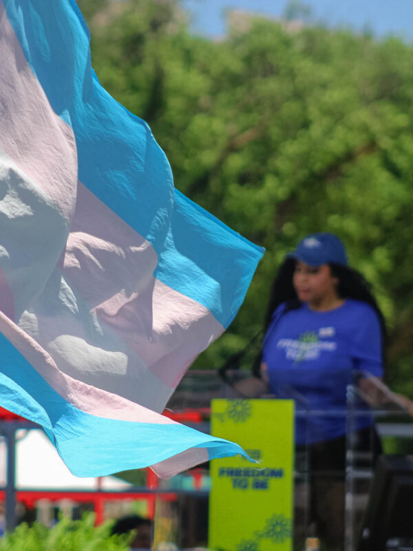 Trans pride flag with a woman speaking on stage in the background.