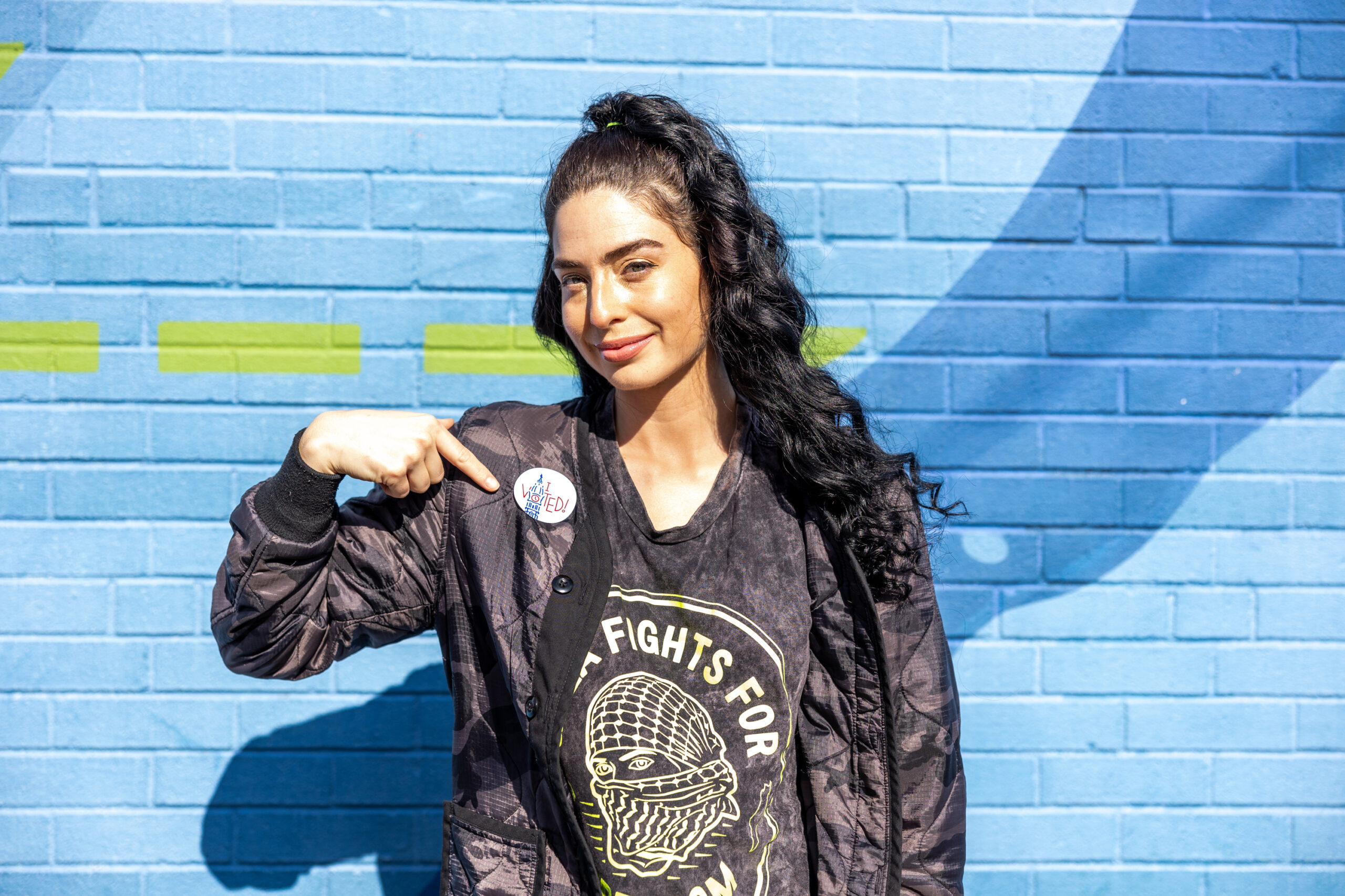 A picture of a woman with long black hair in a high-low ponytail in a black blazer jacket pointing to a voting sticker on a light blue background.