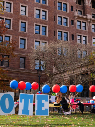 Signs that spell out vote on the grass in front of a large brown brick building that is surrounded by red and blue balloons.