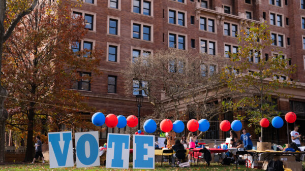 Signs that spell out vote on the grass in front of a large brown brick building that is surrounded by red and blue balloons.