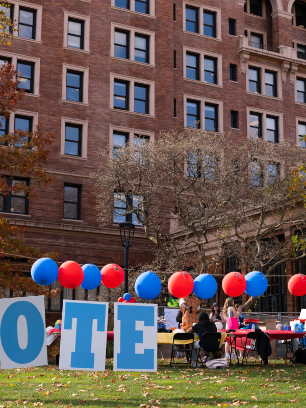Signs that spell out vote on the grass in front of a large brown brick building that is surrounded by red and blue balloons.