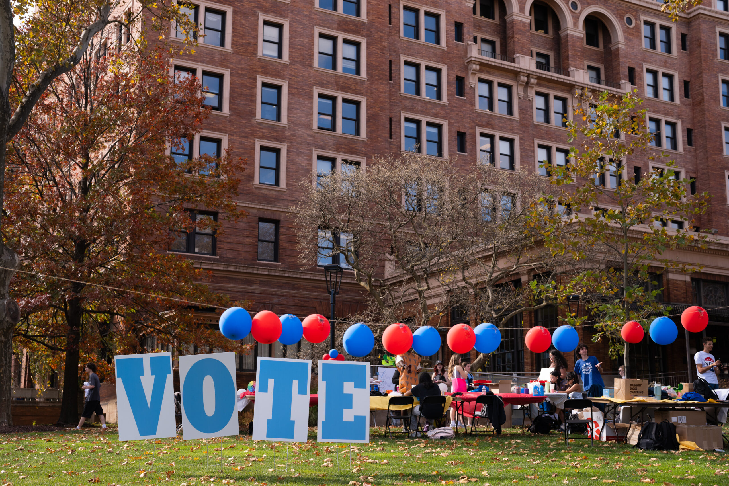 Signs that spell out vote on the grass in front of a large brown brick building that is surrounded by red and blue balloons.