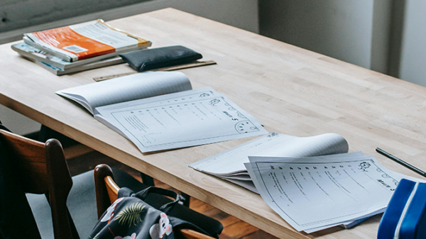 Open books on a wood desk with chairs on a green and white backpack.