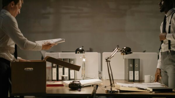 Two men, dressed in a white button down and dark colored slacks, standing across from each other by two desks