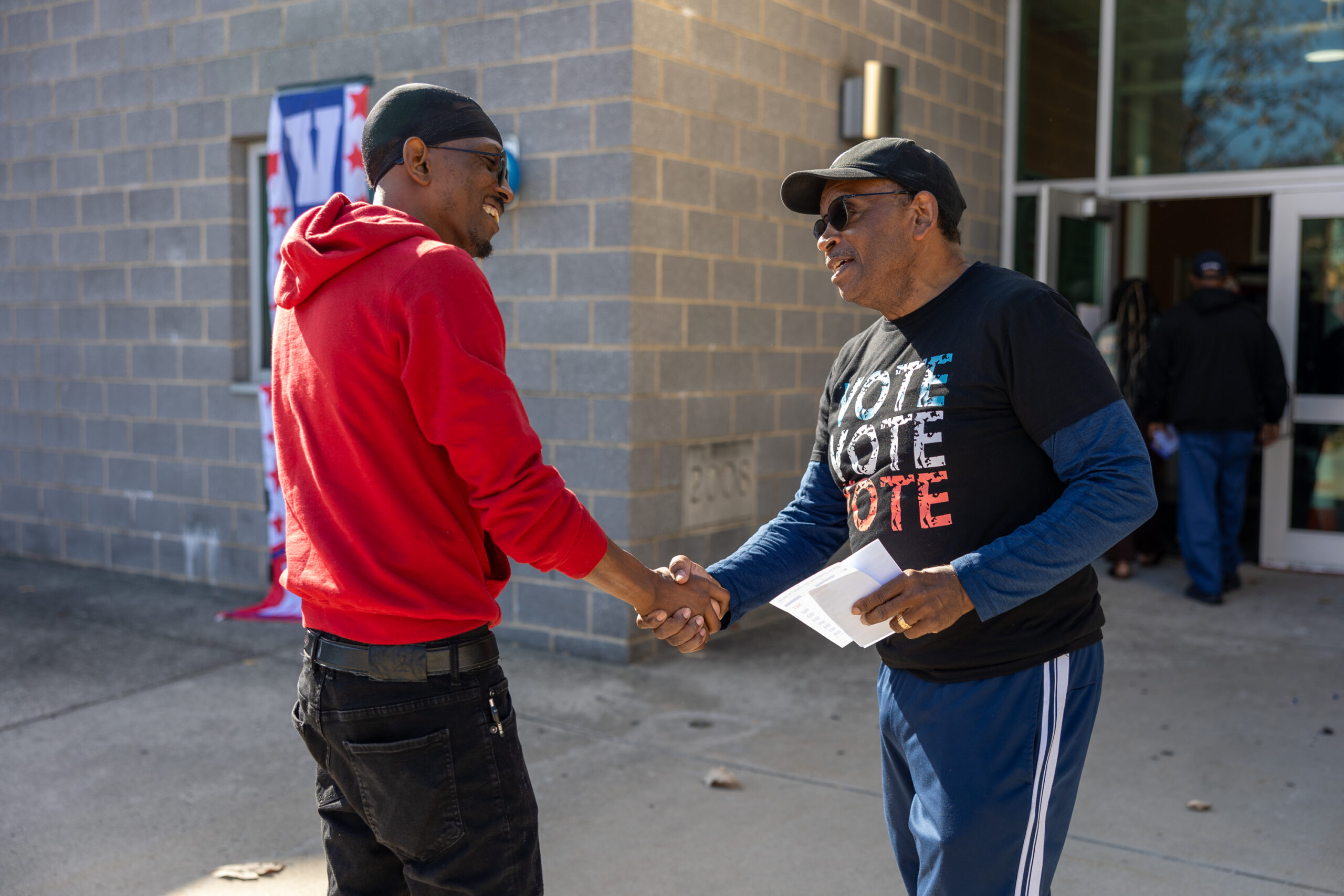 Two people, both in hats, one in a red hoodie and one with a vote shirt on