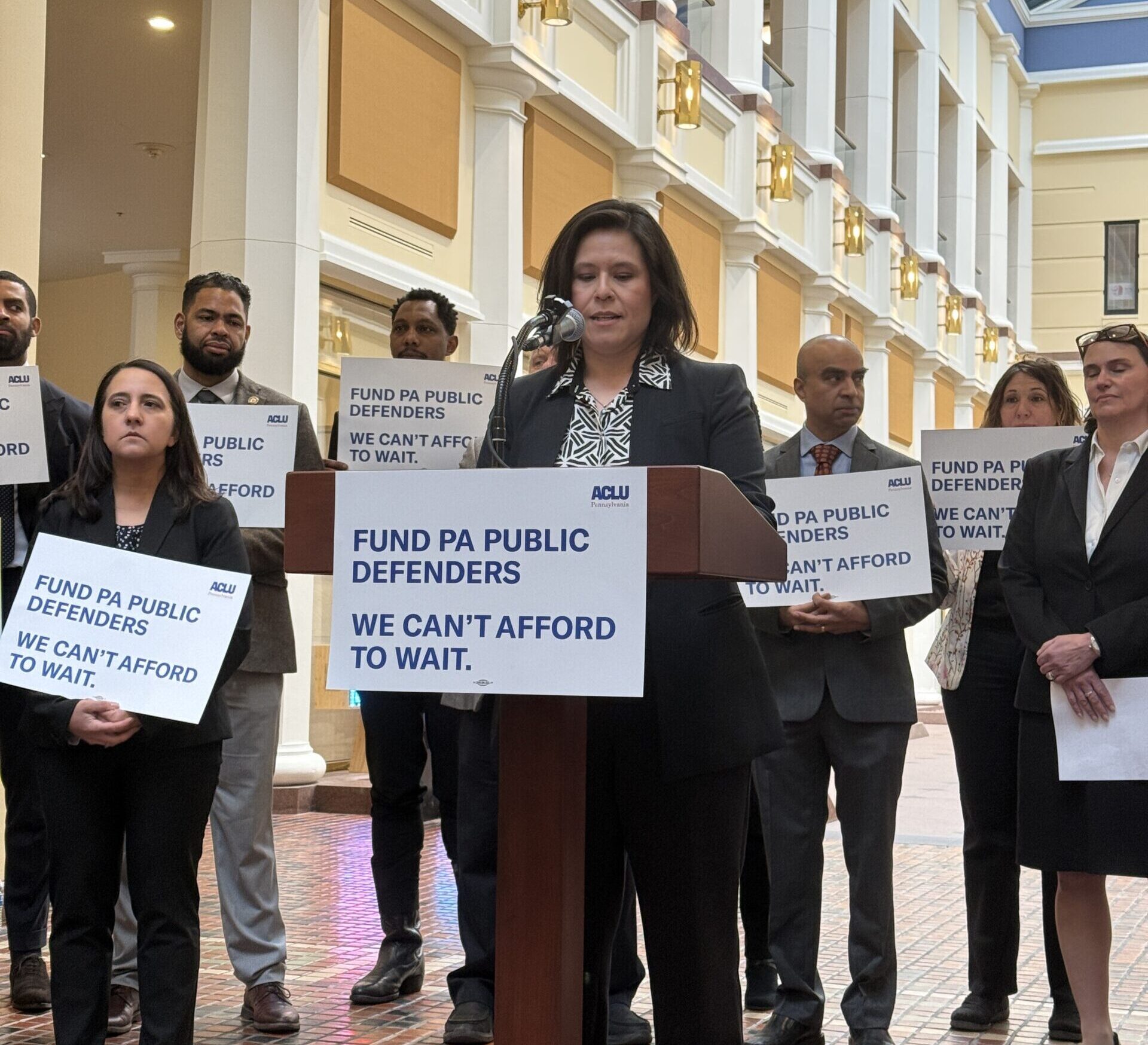 Photo of a group standing at a podium speaking about public defense in PA and holding signs.