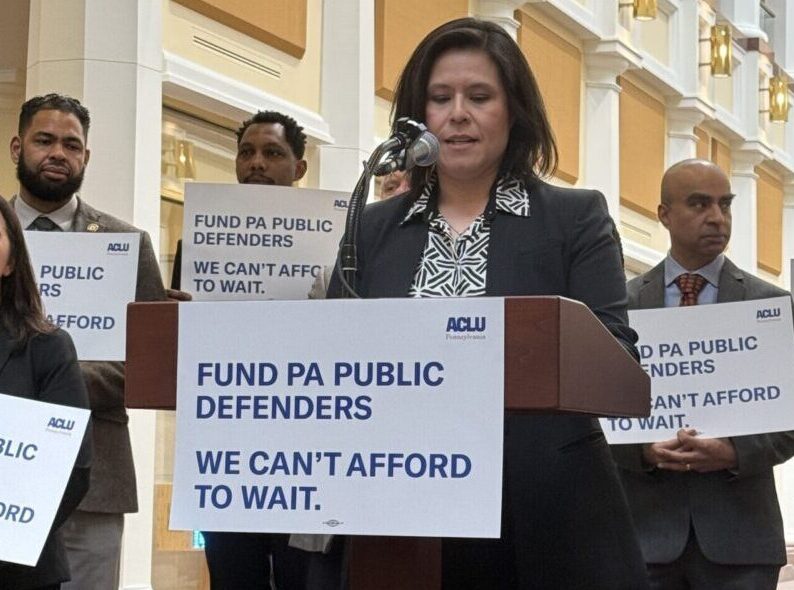 Photo of a group standing at a podium speaking about public defense in PA and holding signs.