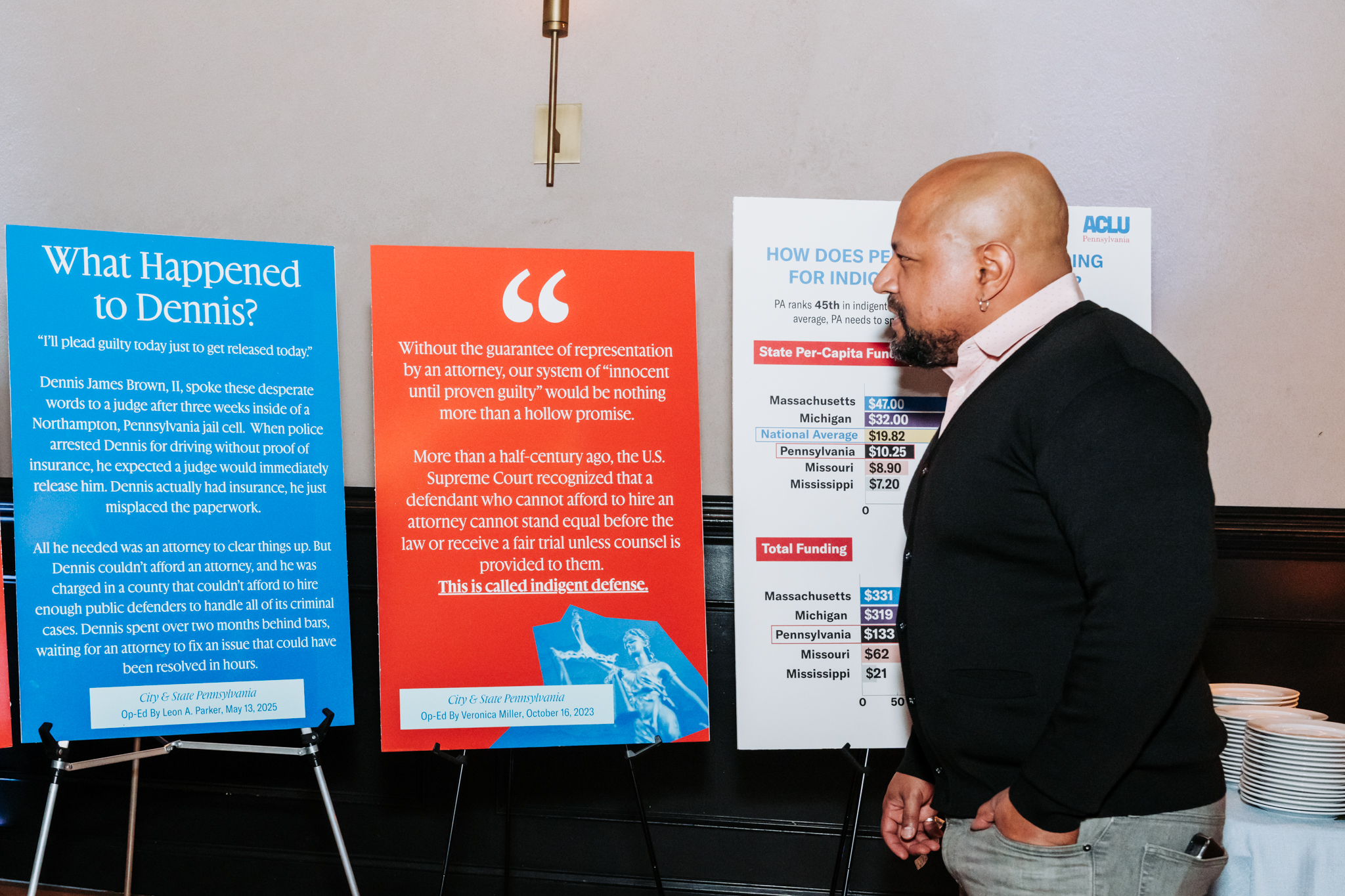 A man with a black cardigan and pink buttoned shirt looking at posters that talk about our public defense work.