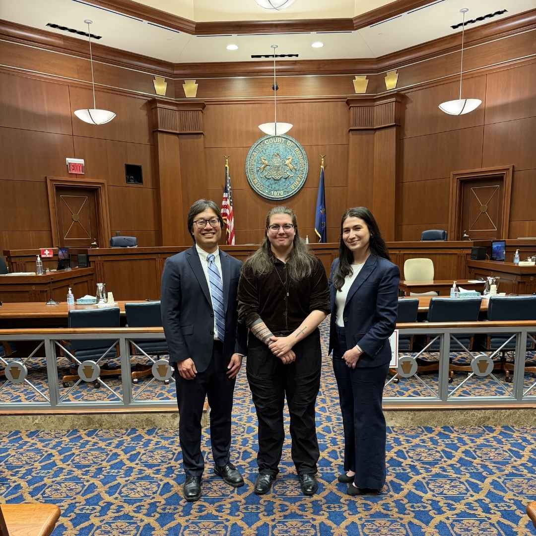 ACLU-PA attorneys stand with our client EJ between them. They are in a courtroom with wooden paneling and a large seal on the wall behind them.