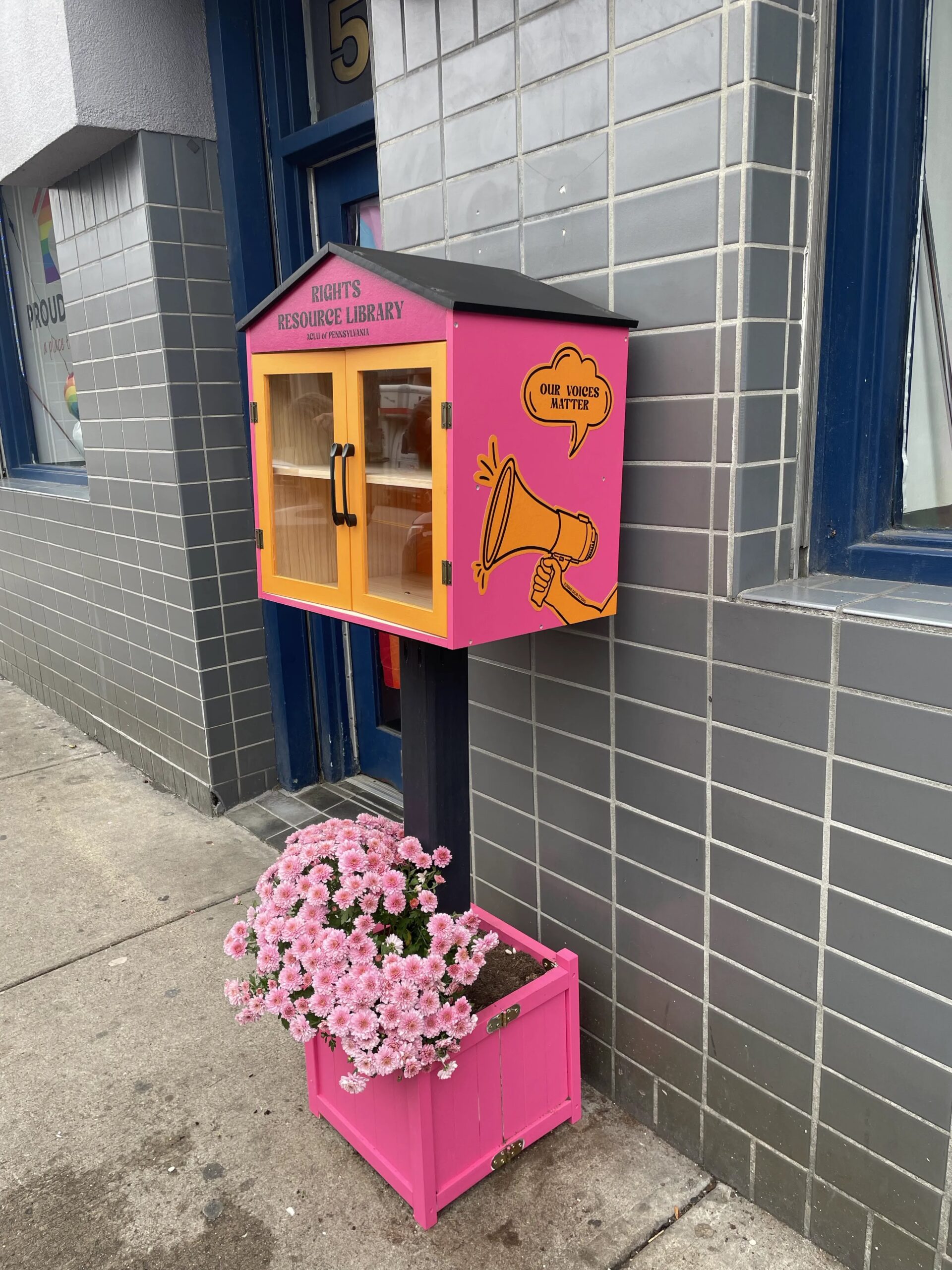 A photo of our Rights Resource Library. The little community library is pink and orange with pink flowers at the base.
