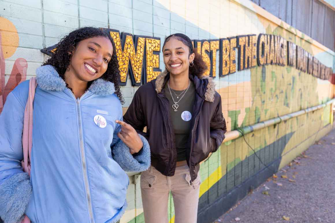Two people, one in a blue puffer jacket and one in a brown coat pointing to their voting stickers on their lapels