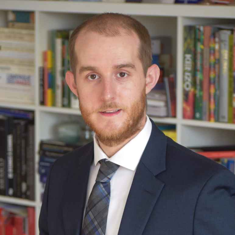 A picture of Keith Armstrong with a beard and navy blue suit with a white button down shirt with a blue plaid tie