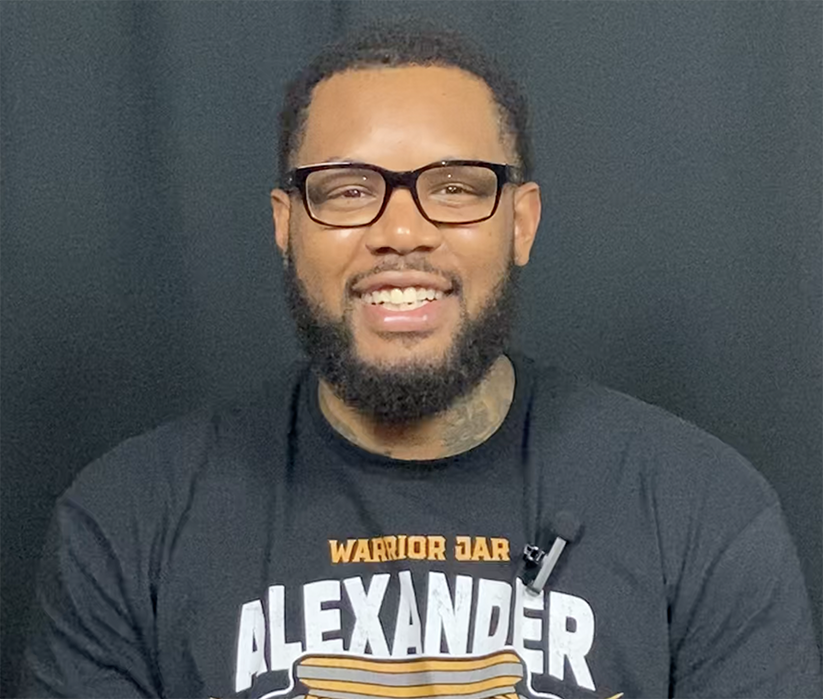 Dontae is smiling while wearing black glasses and a t-shirt that says "Warrior Bar Alexander" on a black background