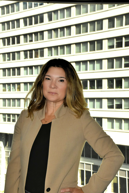 A picture of Rebecca in front of a window with a brown blazer, black shirt, and highlighted brunette and blonde hair.