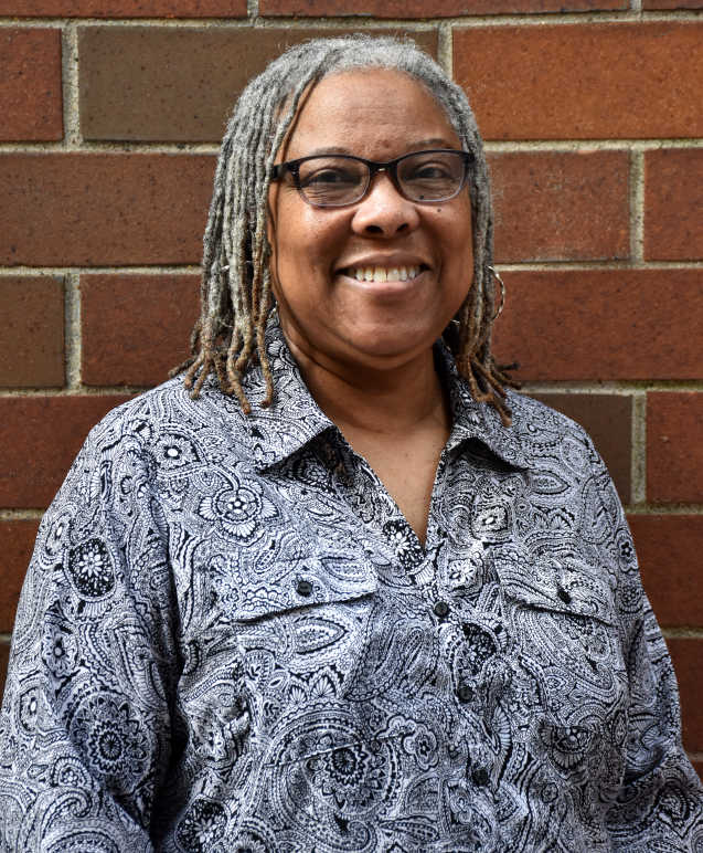 Brenda Allen smiles in a pattenred button down shirt with glasses and gray locs