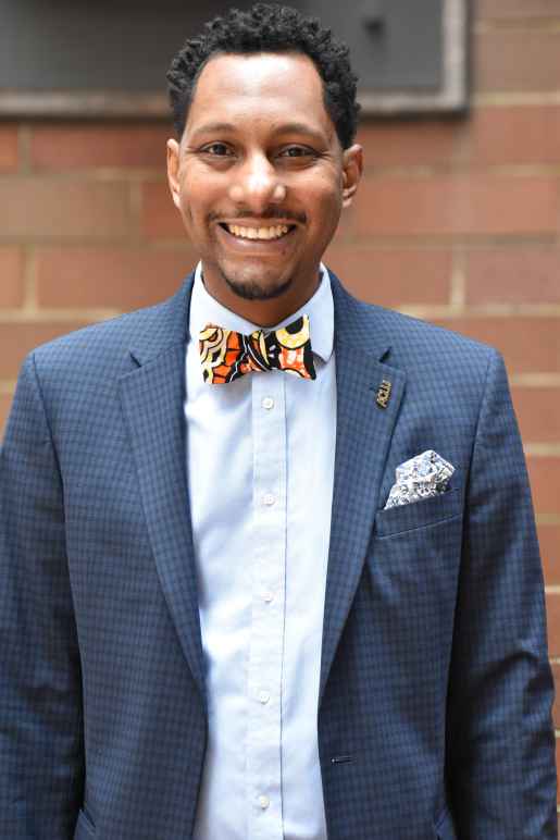 A photo of Mike Lee, smiling, with a light blue button down and dark blue suit blazer with a kente cloth bowtie.